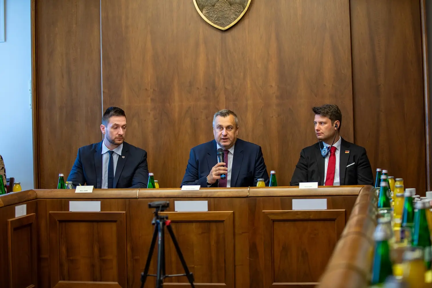 Three speakers at a press conference in a wood-panelled hall, the man in the middle speaking into a microphone