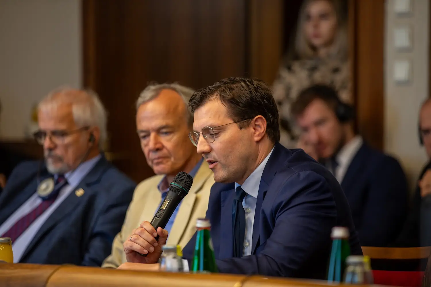 Speaker with a handheld microphone addressing the room, two colleagues seated beside him