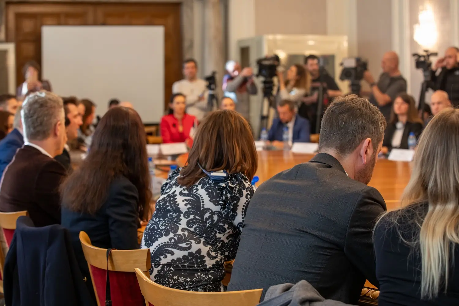 Audience and camera crews at a press event in a historic hall, seen from the back of the room