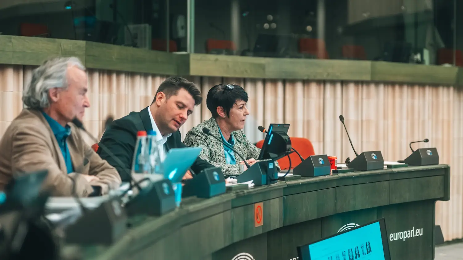 Three participants working side by side at the chair's desk of a European Parliament meeting room